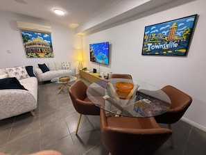 A modern living room with a glass dining table, leather chairs, a sofa, and two wall-mounted paintings. - Oasis Apartments Townsville (Garbutt)