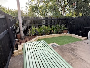 A backyard with a green artificial turf, a wooden bench, and a black fence. - Oasis Apartments Townsville (Garbutt)