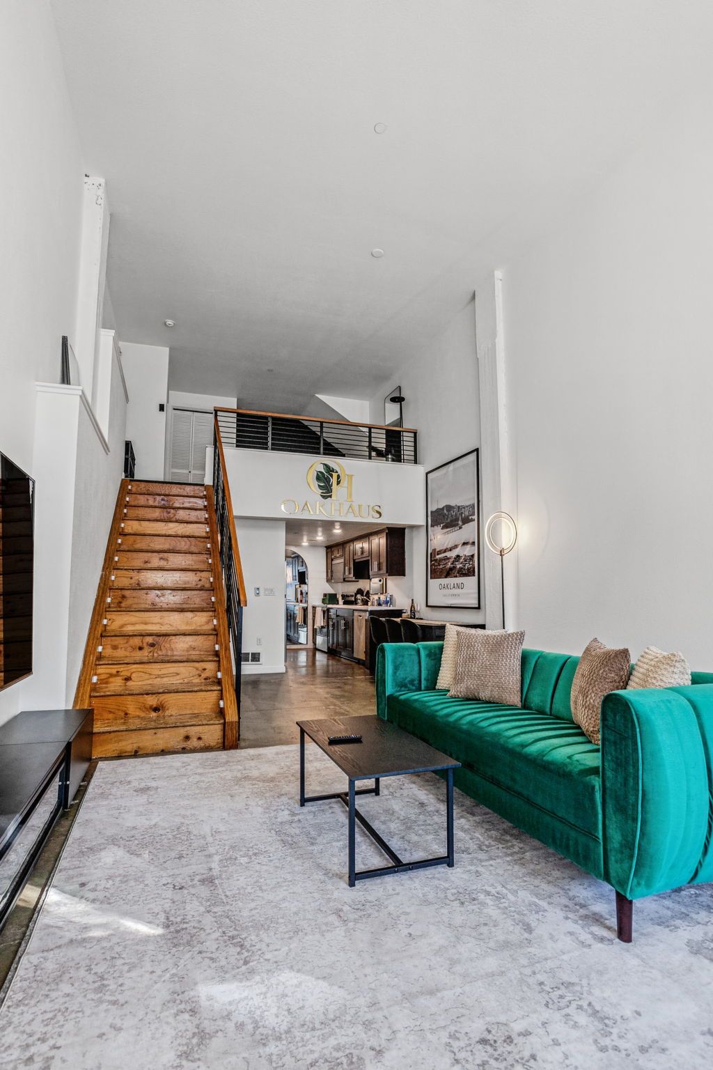 A spacious living area with a wooden staircase, a green sofa, and a kitchen in the background.