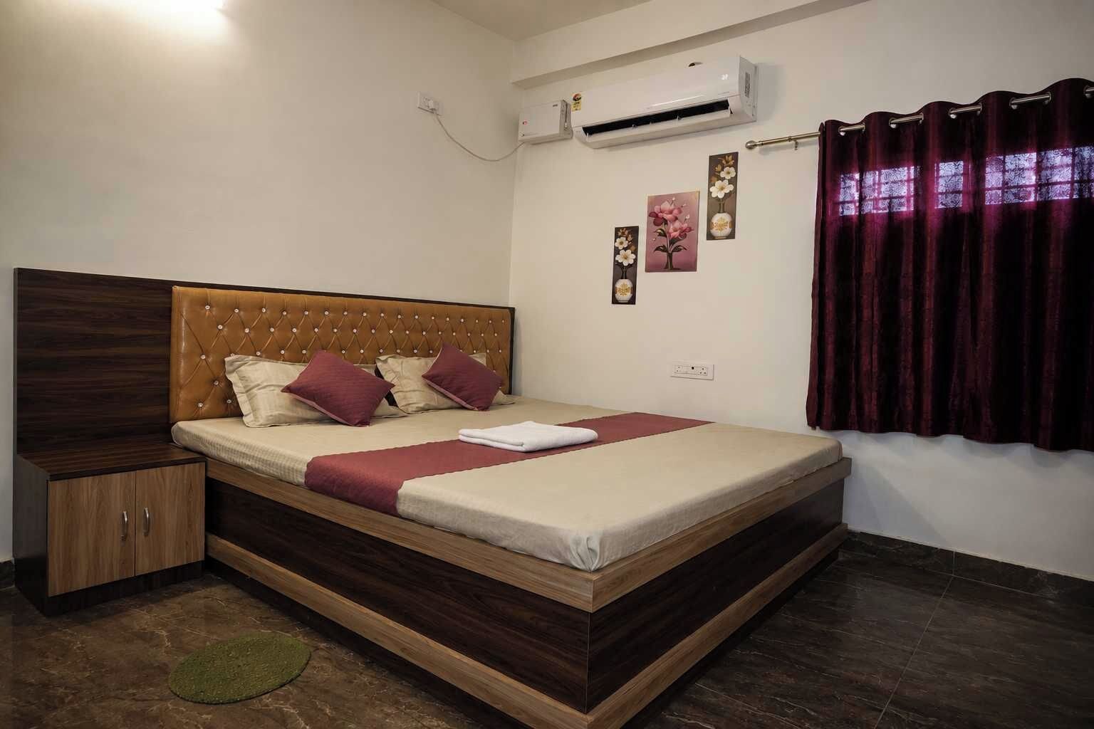 Un dormitorio con cabecera de madera, cama con colcha color beige y borgoña, mesita de noche de madera, alfombra verde, equipo de aire acondicionado y ventana con cortinas moradas.