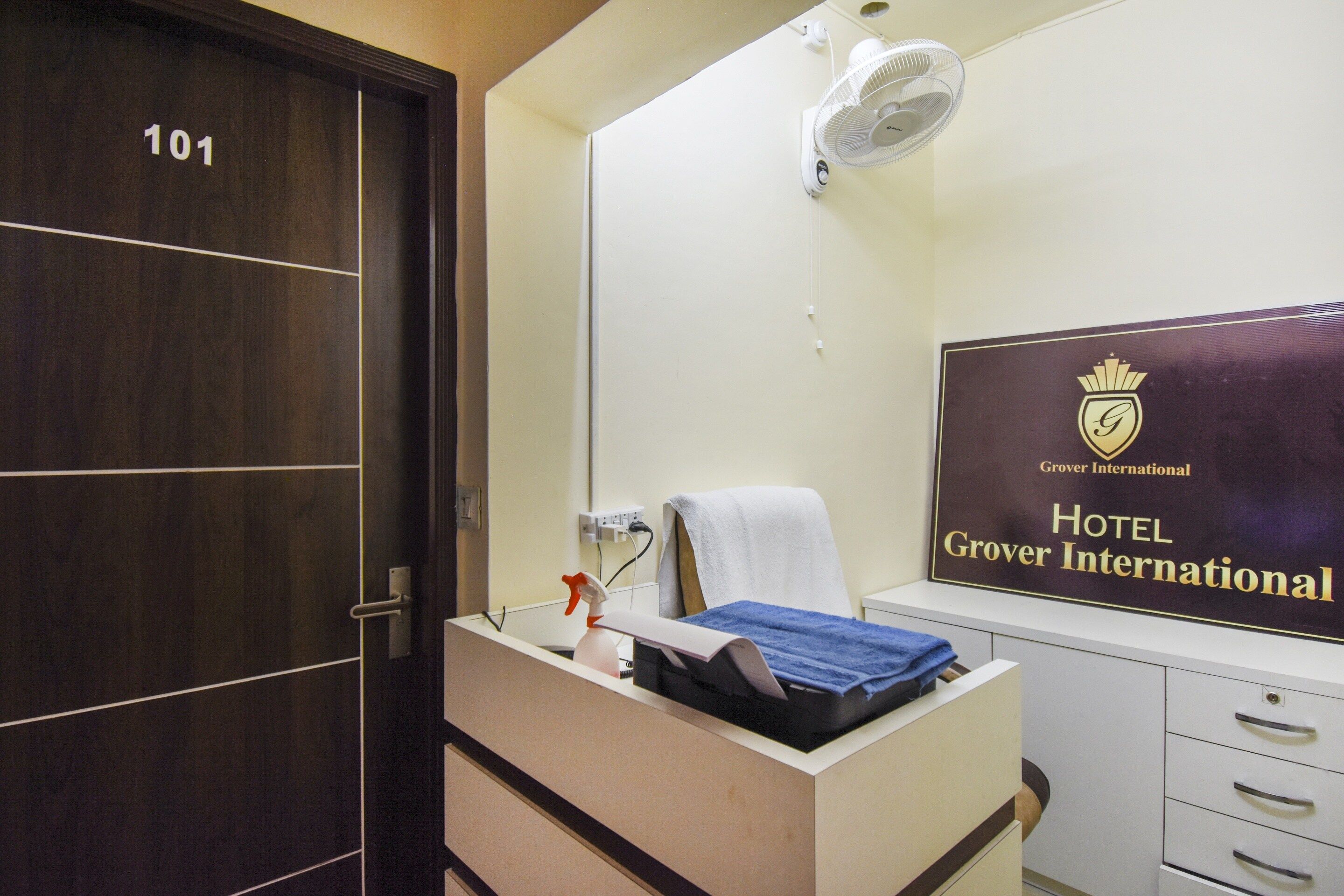 A hotel room with a wooden door labeled '101', a bedside table with a towel and a spray bottle, a wall-mounted fan, and a sign for 'Hotel Grover International'.