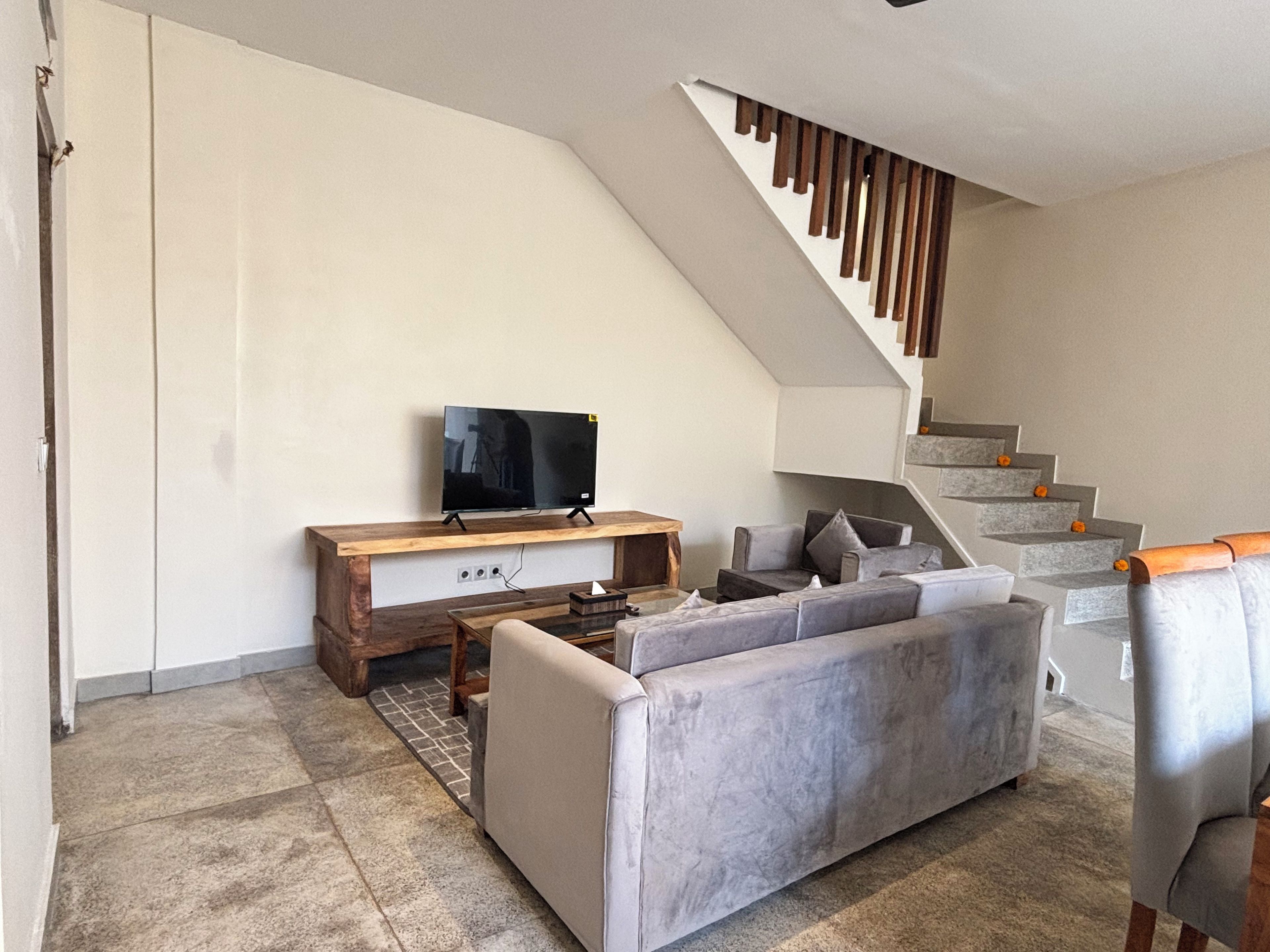 A modern living room with a grey sofa, a wooden coffee table, a flat-screen TV on a wooden stand, and a staircase with a wooden railing.