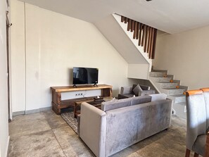 Una sala de estar moderna con un sofá gris, una mesa de centro de madera, un televisor de pantalla plana sobre un soporte de madera y una escalera con barandilla de madera.