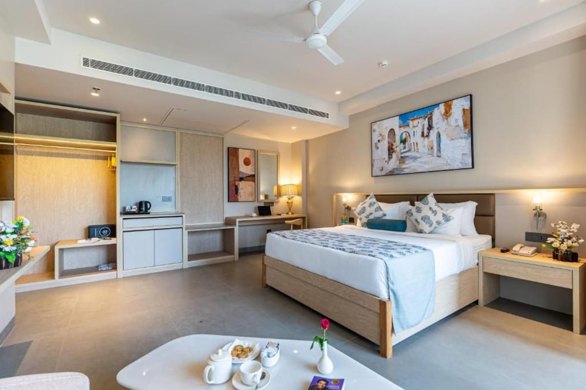 A modern hotel room with a bed, bedside table, desk, and a kitchenette.