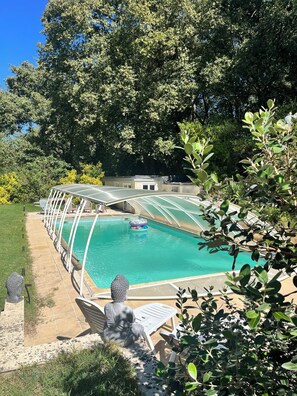 Una piscina con cerramiento de vidrio, rodeada de follaje y una estatua.