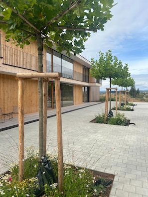 Een modern gebouw met een houten gevel, terrasplekken en een bestraat binnenterrein met planten en bomen.