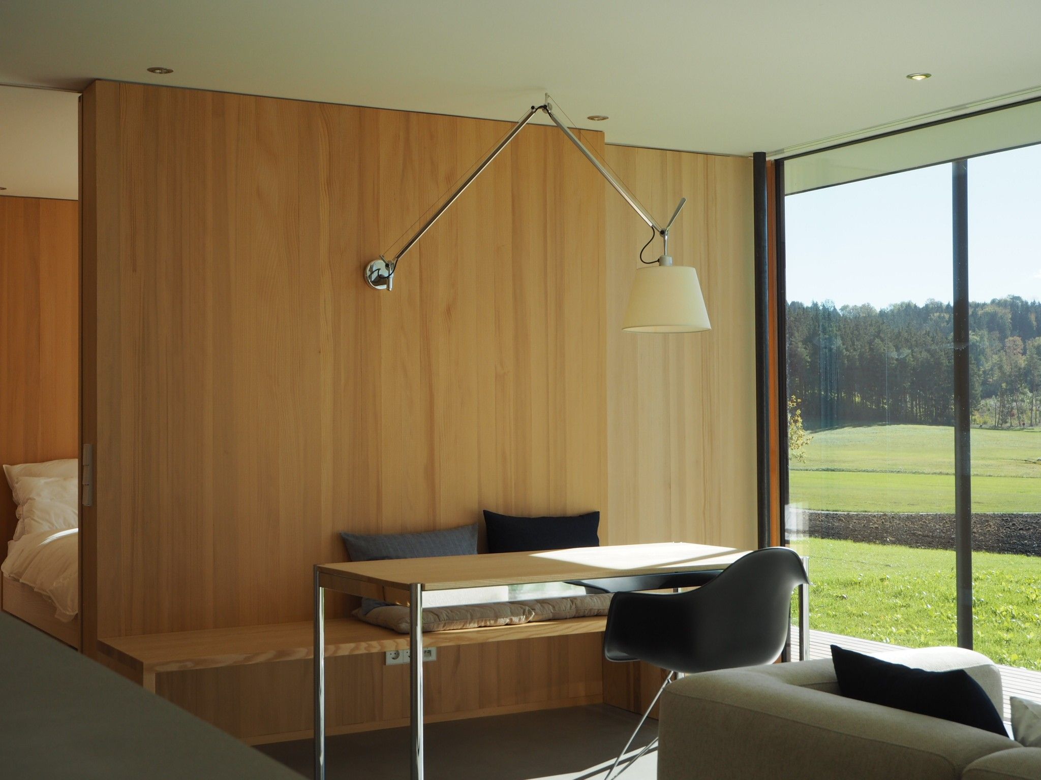 Ein modernes Zimmer mit einer Holzwand, einem Bett, einem Schreibtisch mit Stuhl und einem großen Fenster mit Blick auf eine grüne Landschaft.