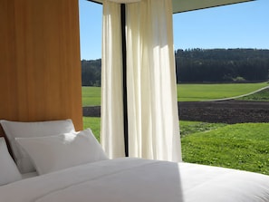 Een slaapkamer met een groot bed, witte lakens en uitzicht op een groen veld en bos door dunne gordijnen.
