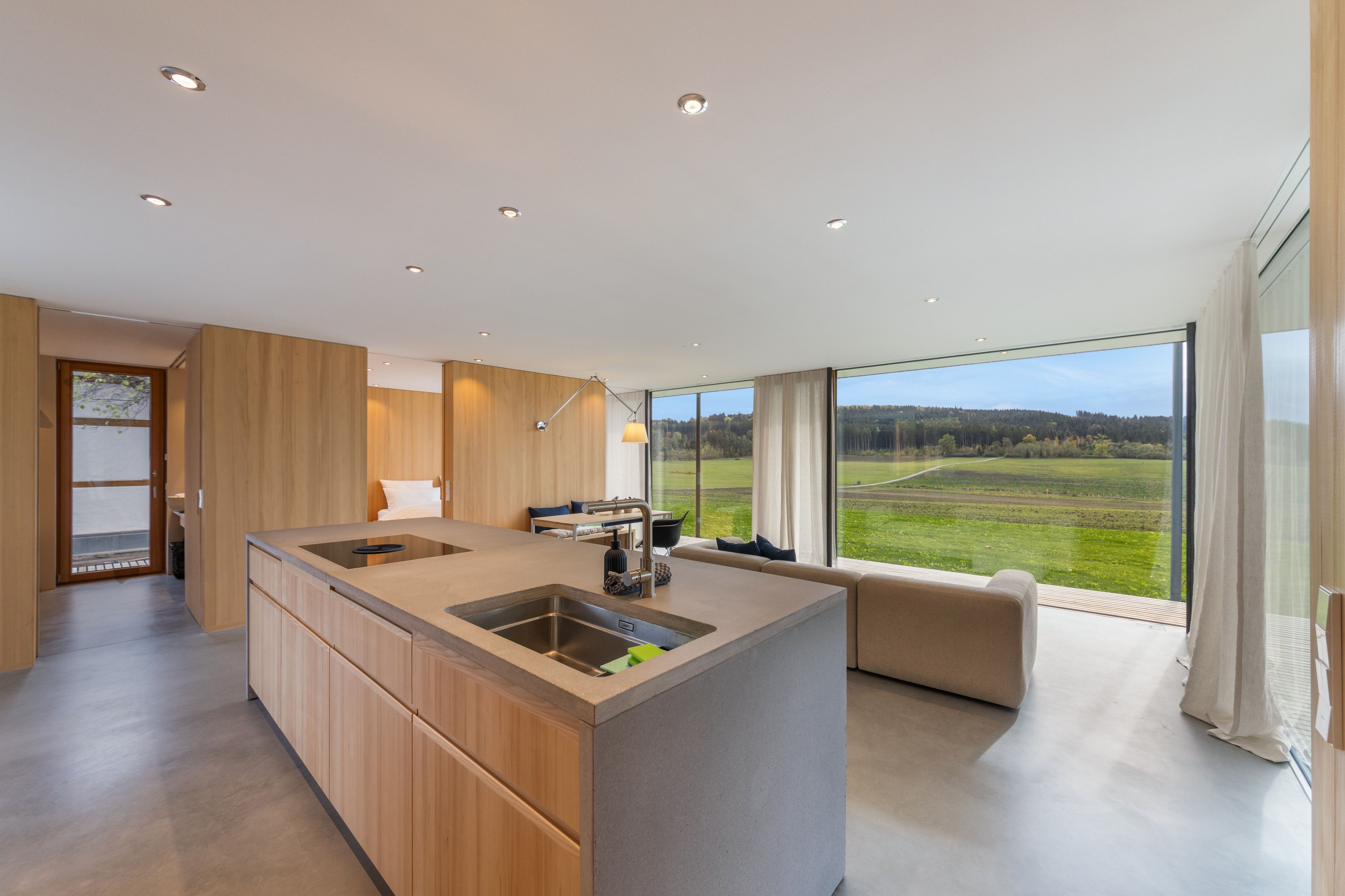 Eine moderne Küche mit einer großen Kochinsel, einem Essbereich mit einer Couch und Blick auf eine grüne Landschaft durch große Fenster.