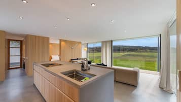 Eine moderne Küche mit einer großen Kochinsel, einem Essbereich mit einer Couch und Blick auf eine grüne Landschaft durch große Fenster.