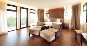 Quarto de hotel espaçoso com uma cama grande, dois sofás, uma mesa pequena e vista para o verde através de grandes janelas.