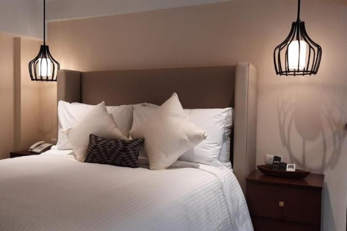 Quarto de hotel com cama, mesas de cabeceira e luminárias pendentes.