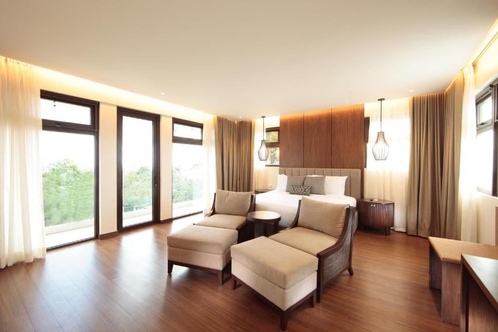 Habitación de hotel con una cama grande, dos sofás, una mesita y vistas a zonas verdes a través de las ventanas.