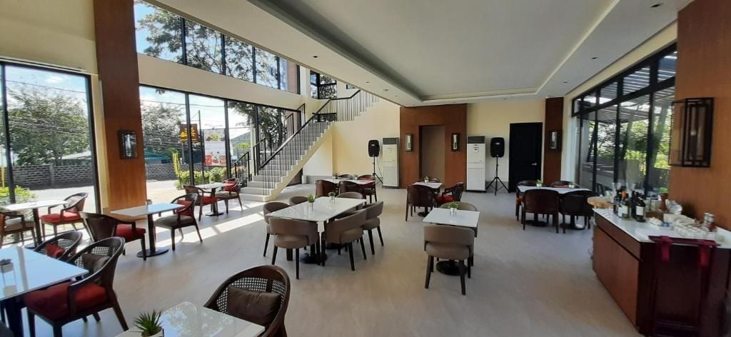Un amplio comedor interior con varias mesas y sillas, ventanas grandes y una escalera que sube a un nivel superior.