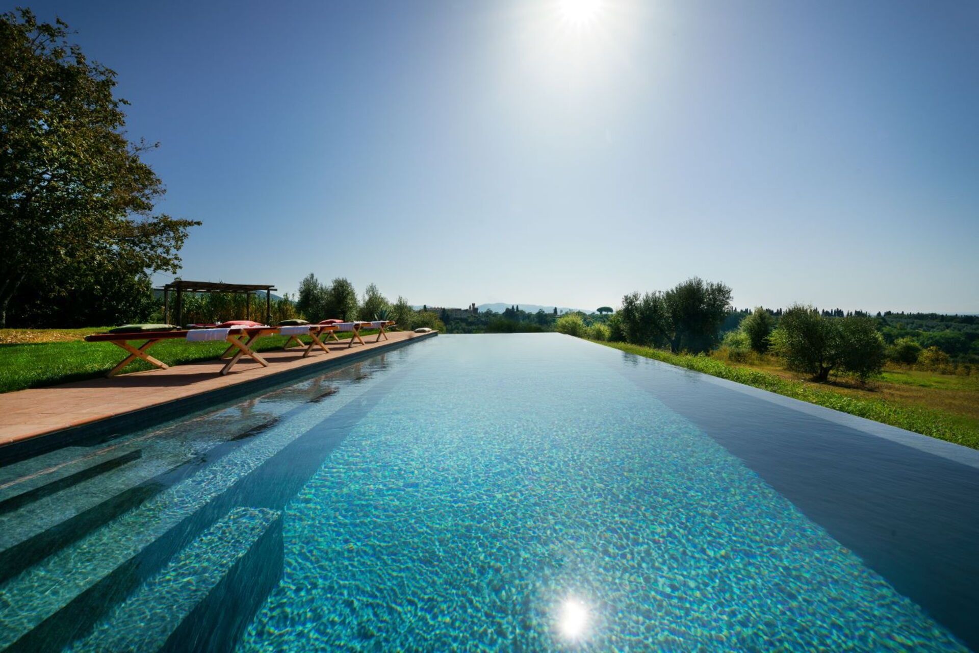 Une piscine aux eaux cristallines, accessible par des marches, entourée de verdure et surplombée d’un ciel dégagé.