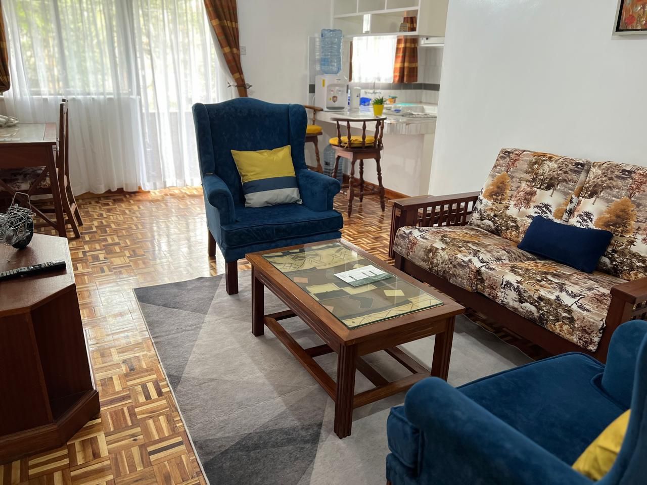 Un salon avec un canapé, un fauteuil et une table basse. On aperçoit un coin cuisine au fond.