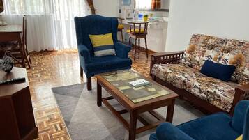 Un salon avec un canapé, un fauteuil et une table basse. On aperçoit un coin cuisine au fond.