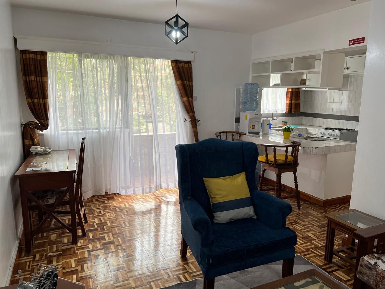 Un salon avec un fauteuil bleu, une table à manger avec des chaises, un coin cuisine avec des placards et une fenêtre avec des rideaux.