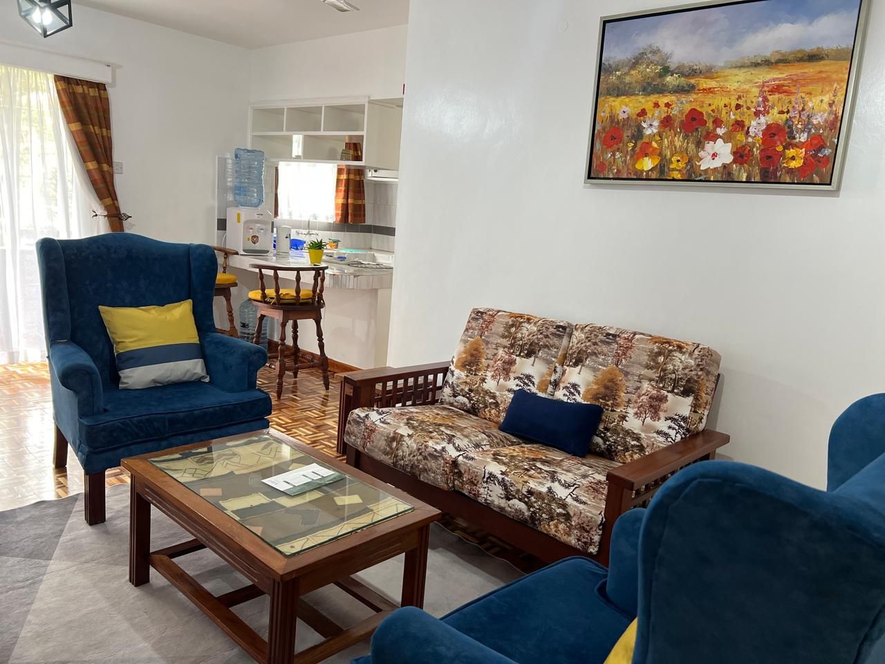 Een woonkamer met een bank, fauteuil en salontafel. Aan de muur hangt een schilderij en er is een keuken op de achtergrond zichtbaar.