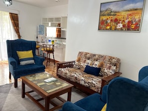 Een woonkamer met een bank, fauteuil en salontafel. Aan de muur hangt een schilderij en er is een keuken op de achtergrond zichtbaar.