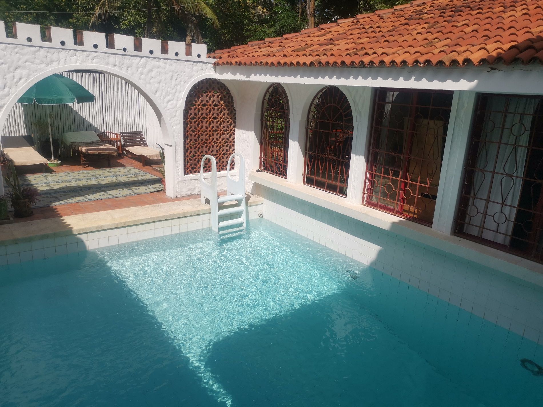 Una piscina con una scala, un'area salotto coperta e un muro bianco con finestre ad arco.
