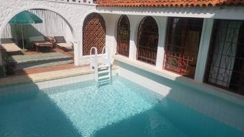 Una piscina con una scala, un'area salotto coperta e un muro bianco con finestre ad arco.