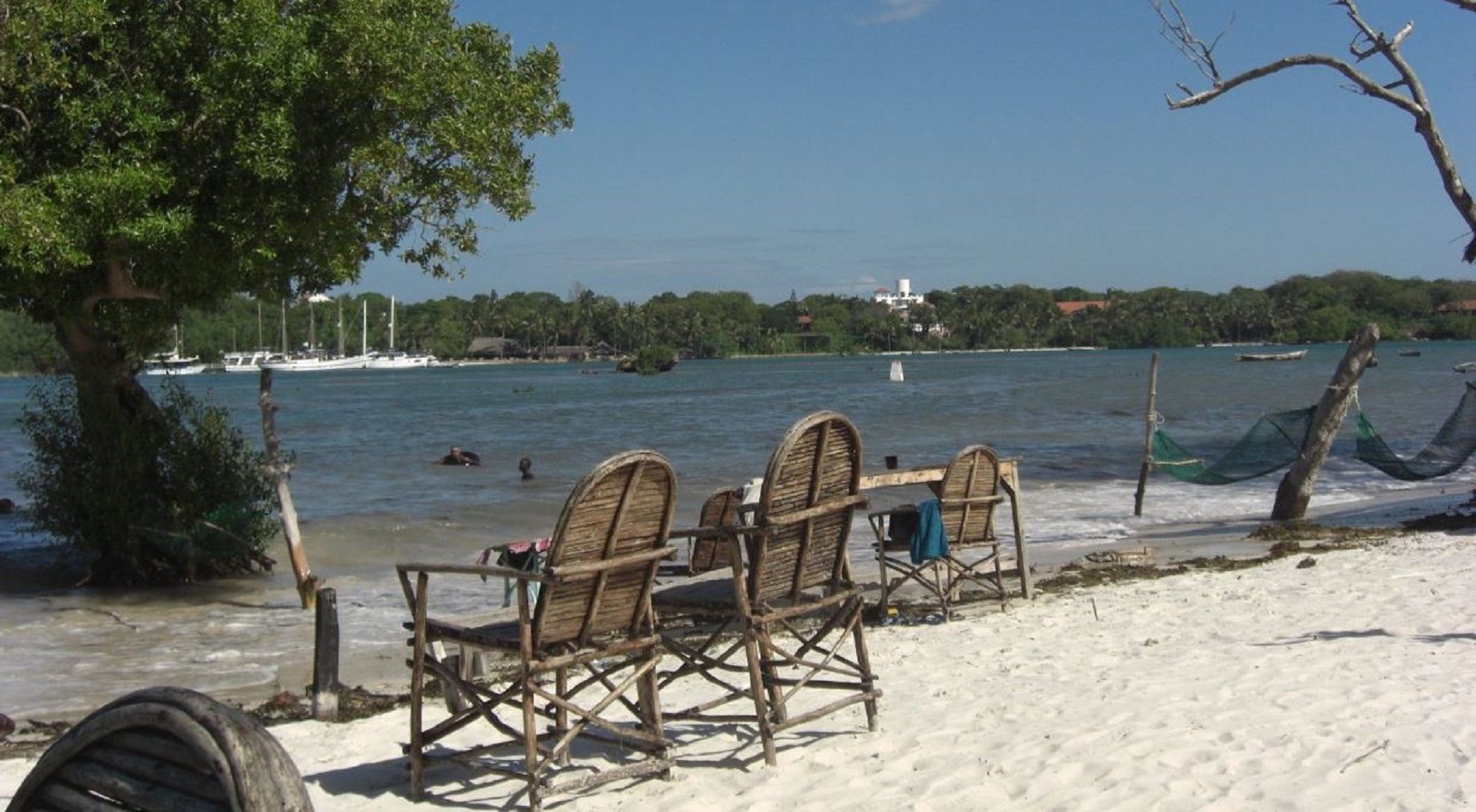 Ein Strand mit Holzstühlen und einem Baum, ruhiges Meer und Boote in der Ferne.