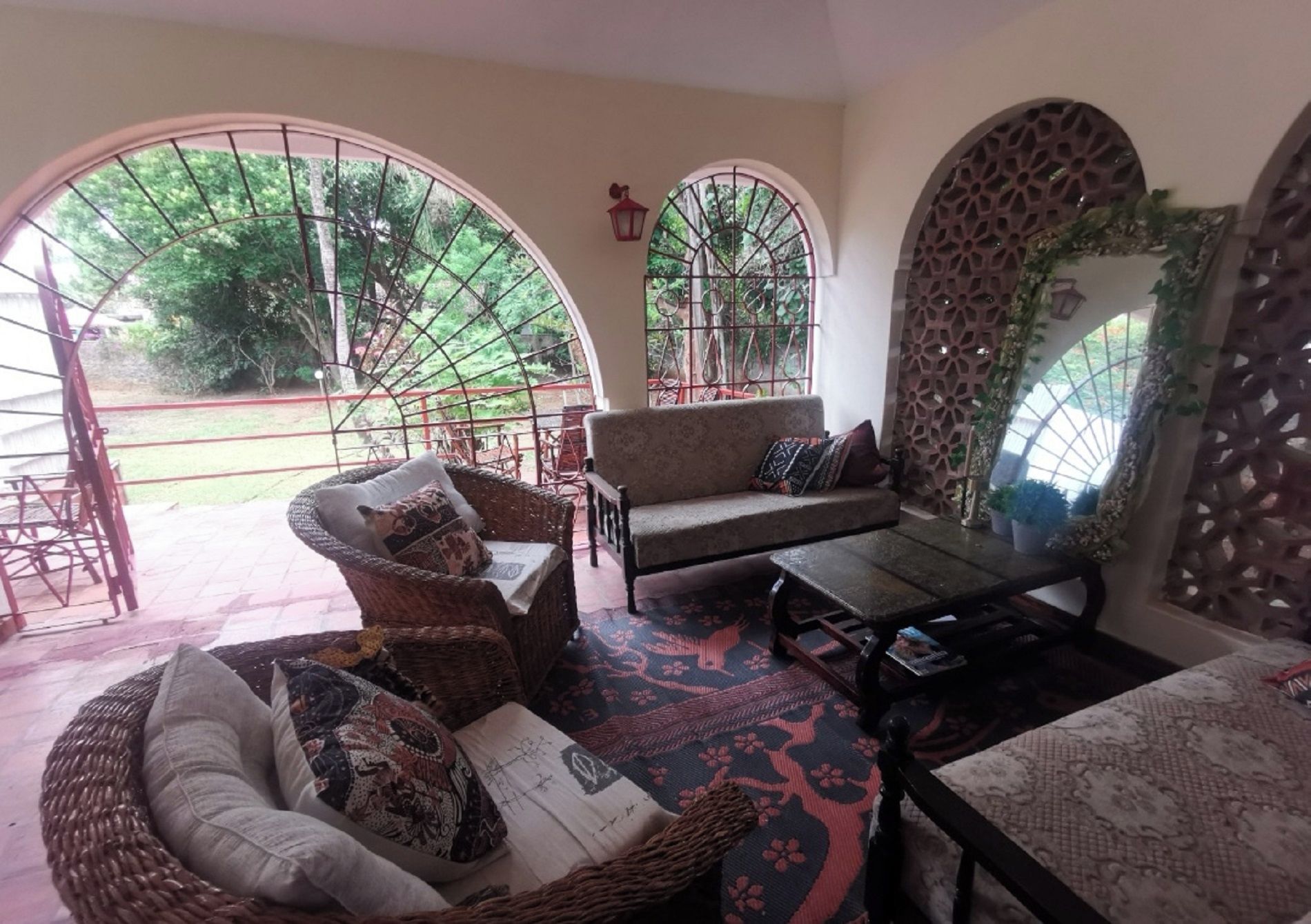 A cozy interior with a sofa, a wicker chair, and a table. There are arched windows and a view of greenery outside.