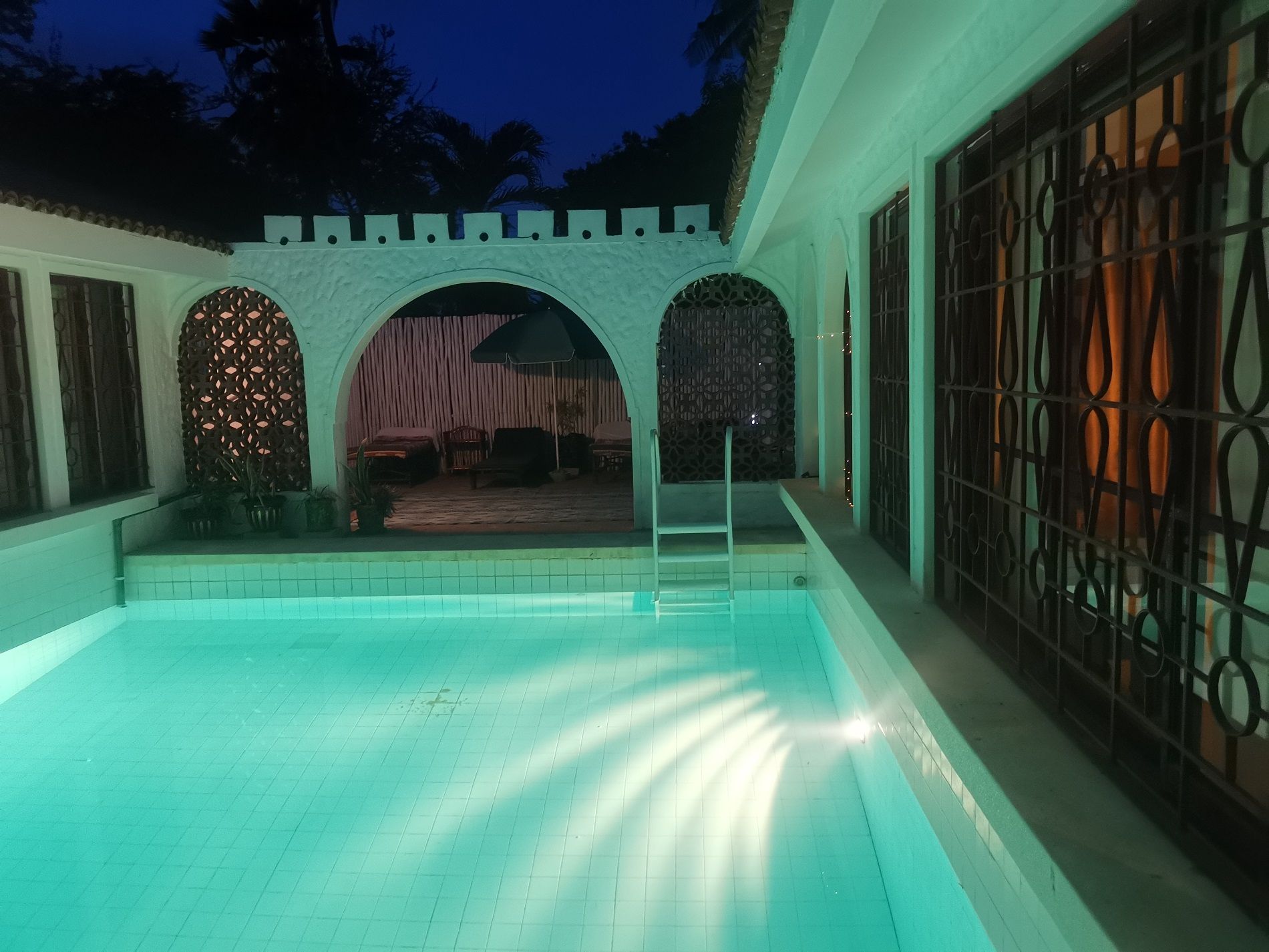 Una piscina con bordo piastrellato, circondata da un portico coperto, una parete in pietra e un cancello in metallo.