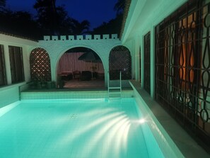 En swimmingpool med fliseterrasse, omgivet af en overdækket patio med stenvæg og metalport.