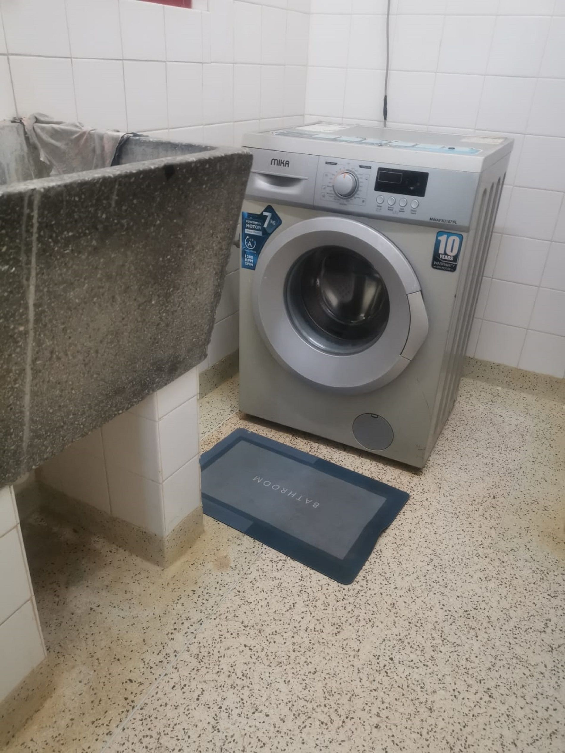 A compact, modern laundry room with a front-loading washing machine, a granite countertop, and a blue mat with 'BATHROOM' written on it.