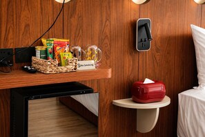 Habitación de hotel con pared de madera, un cartel de 'No Fumar', una cesta de artículos de aseo, un cubo de basura rojo y una mesita de noche con cubrecama blanco.