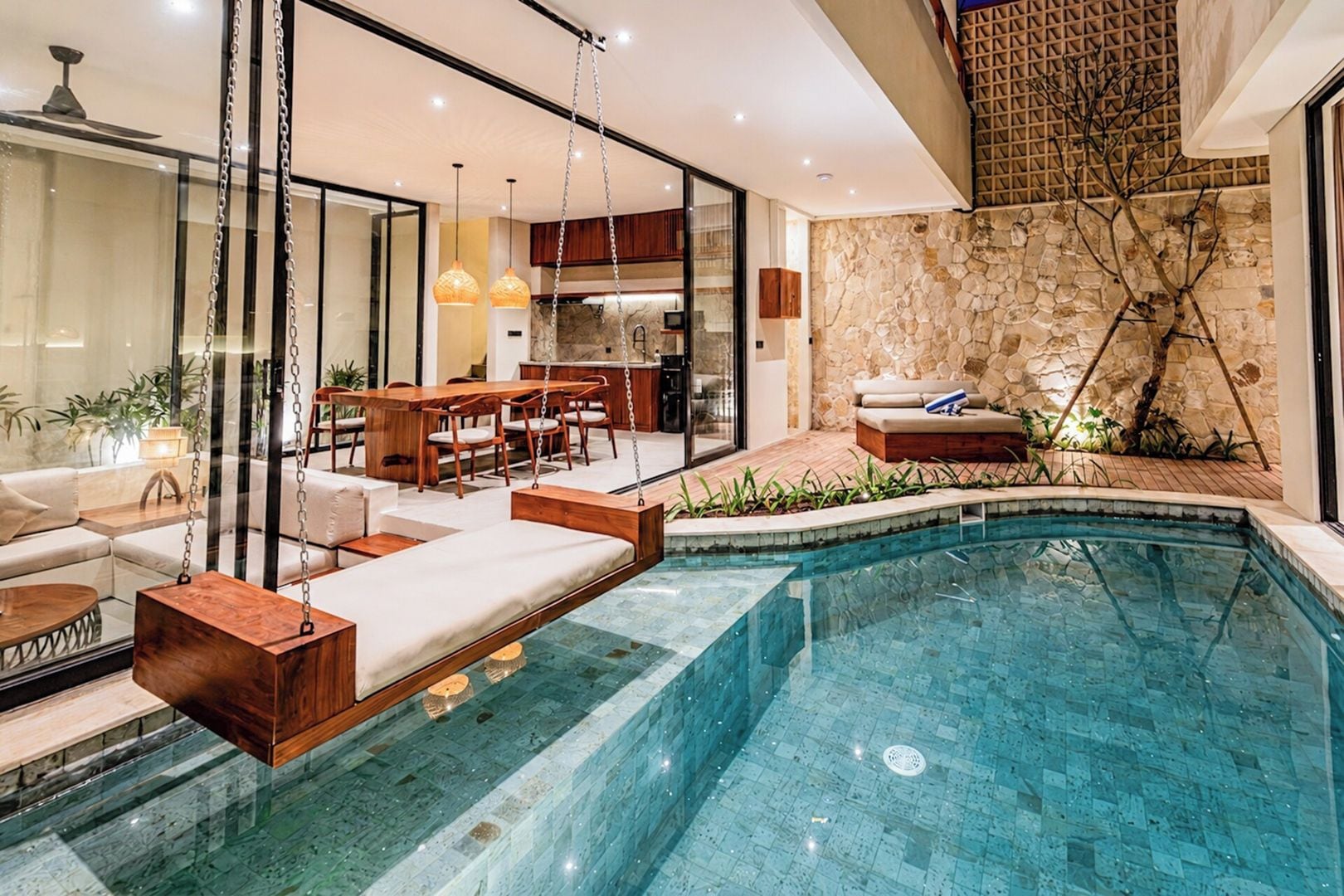A modern indoor swimming pool with a stone wall and hanging chairs.