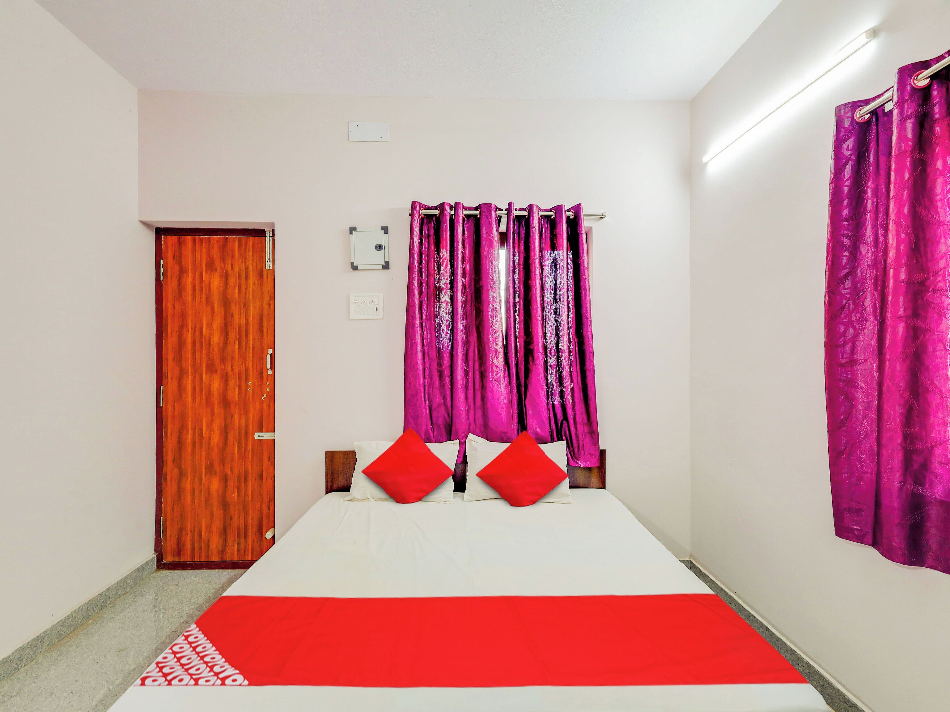 Una habitación con cama, puerta de madera, luz empotrada en la pared y cortina.