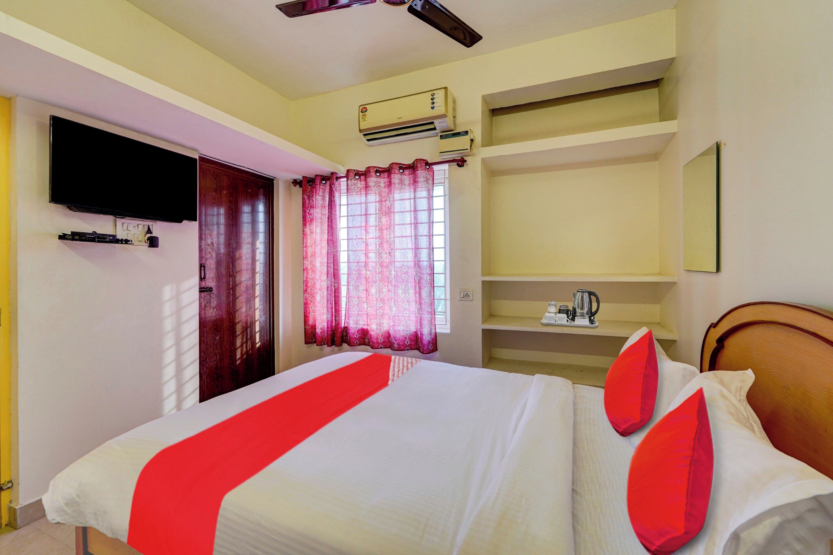 Habitación de hotel con cama, televisor montado en la pared, ventilador de techo, equipo de aire acondicionado, ventana con cortinas y repisa con tetera y tazas.