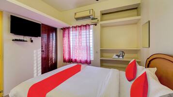 Habitación de hotel con cama, televisor montado en la pared, ventilador de techo, equipo de aire acondicionado, ventana con cortinas y repisa con tetera y tazas.