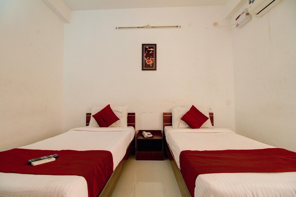 Quarto de hotel com duas camas de solteiro, cada uma com manta e travesseiro vermelhos, uma mesinha de cabeceira com telefone, e um quadro na parede.