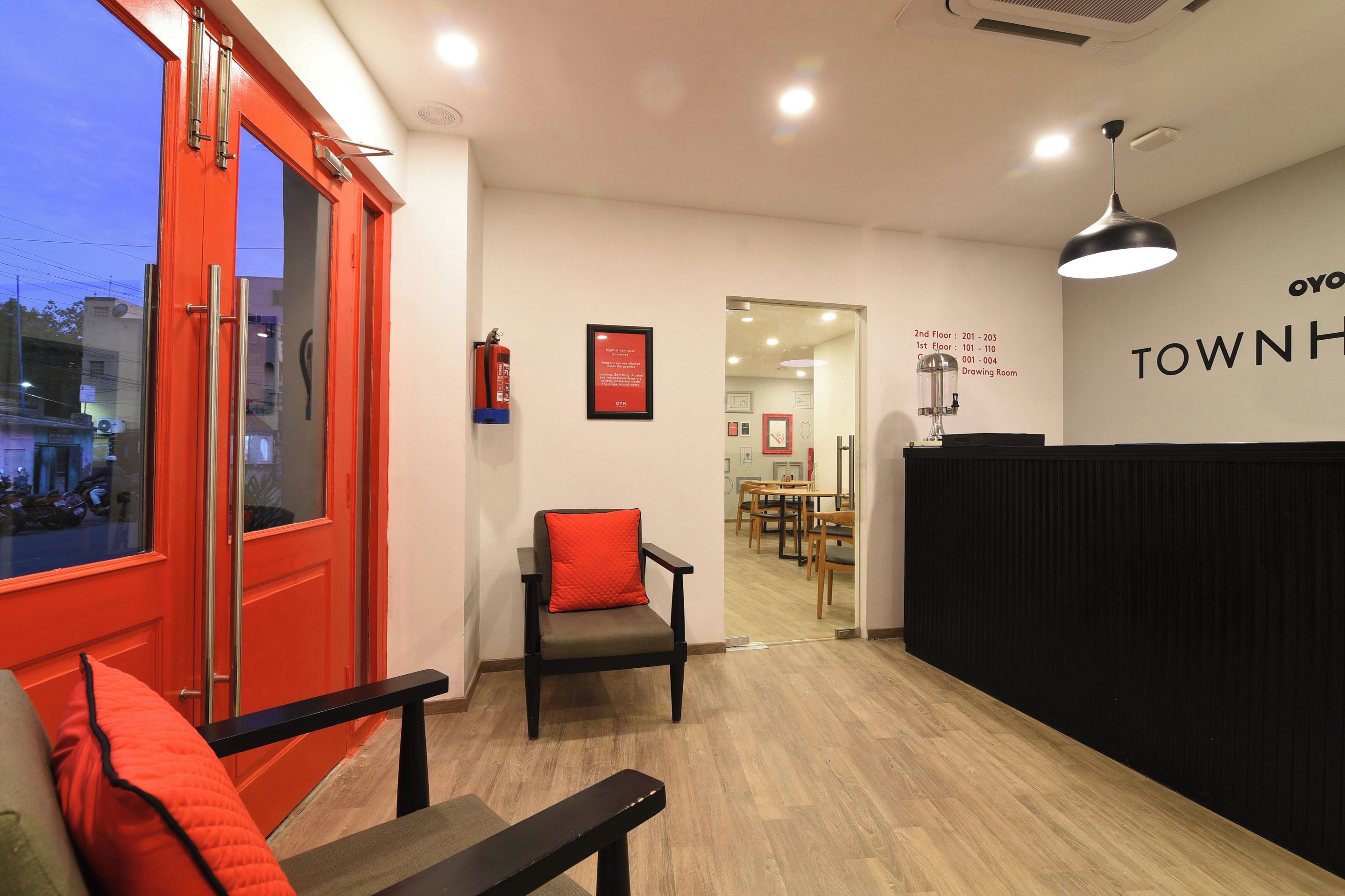 Un área de recepción con una puerta roja, un banco con cojín rojo, un extintor y una pared con el texto 'TOWNH'.