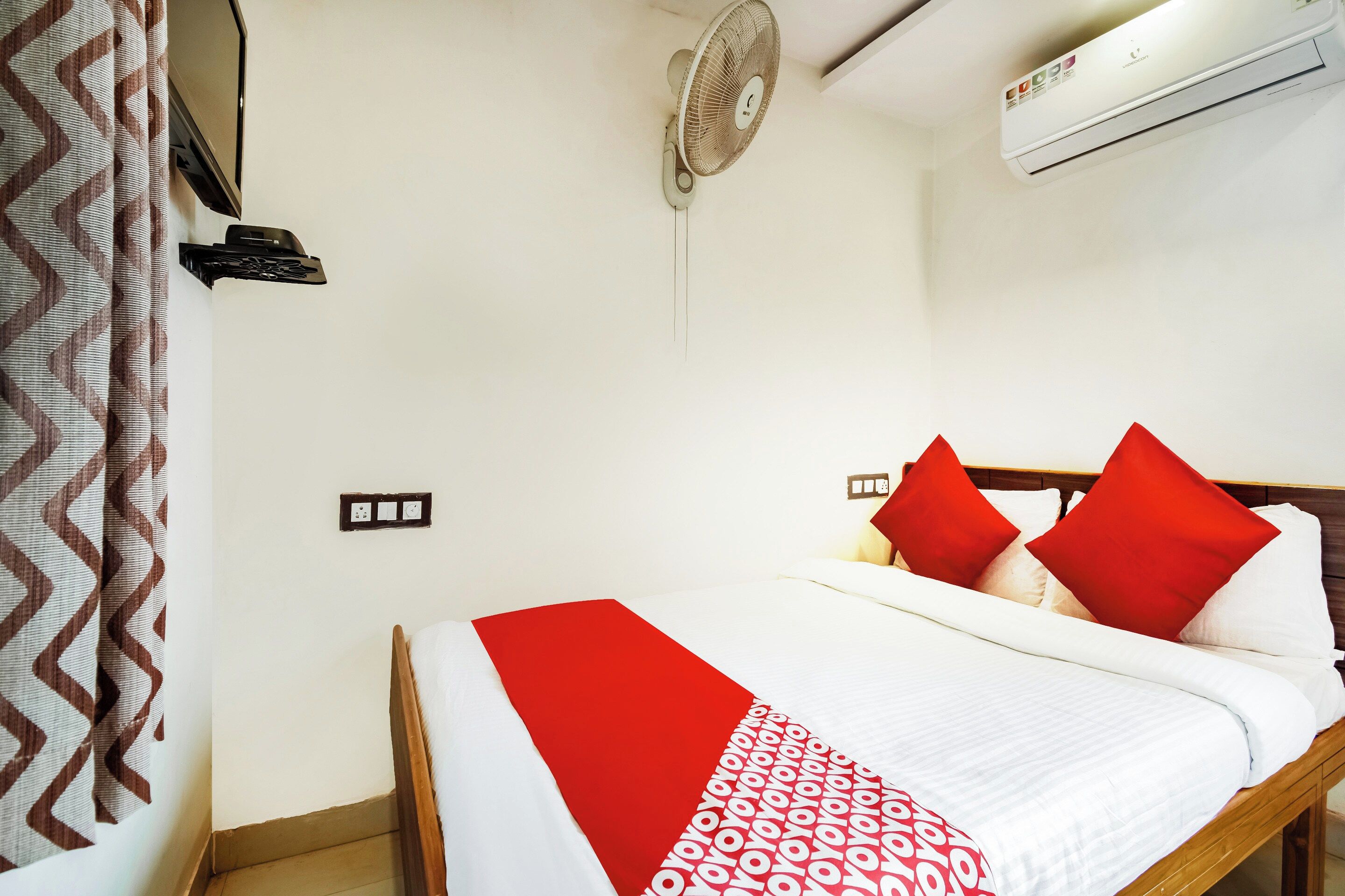 A hotel room with a bed, red pillows, a white and red blanket, a standing fan, an air conditioning unit, and a wall-mounted telephone.