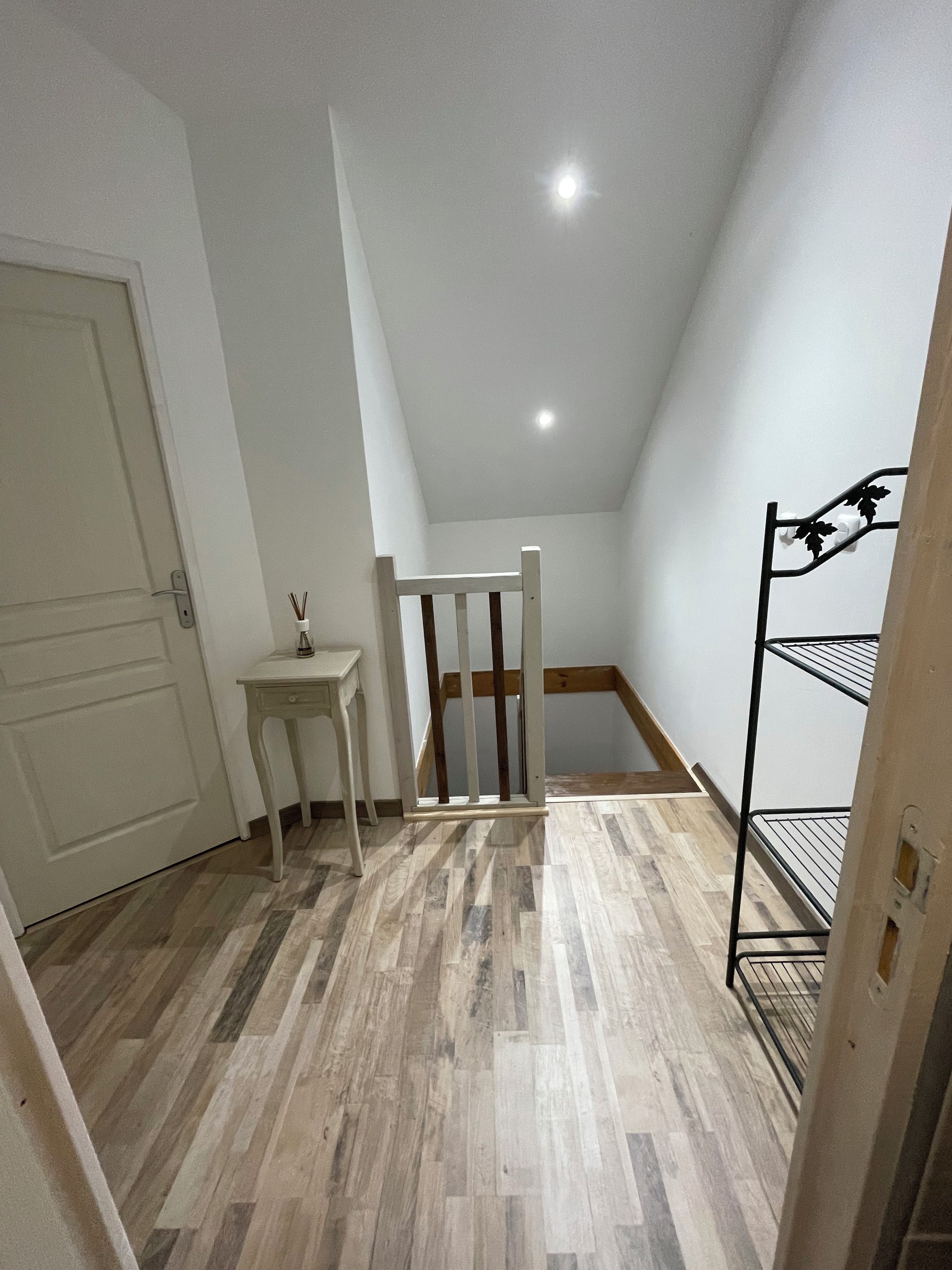 A narrow hallway with a white door, a small table with a candle, a wooden chair, and a metal shelf unit.