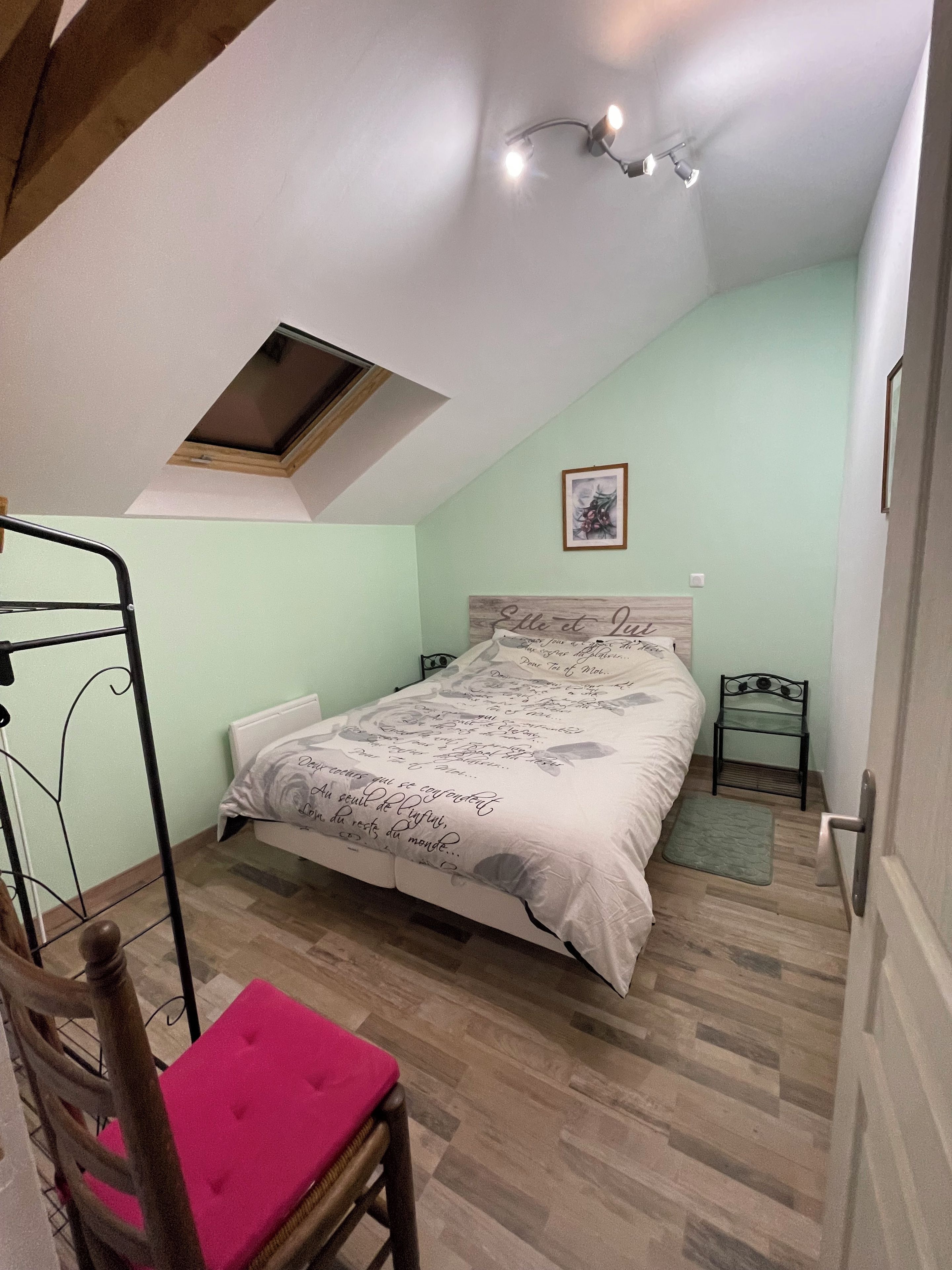 Een slaapkamer met een schuine kap, dakraam, bed met decoratieve hoofdbord, nachtkastje, stoel met roze kussen en een houten vloer.