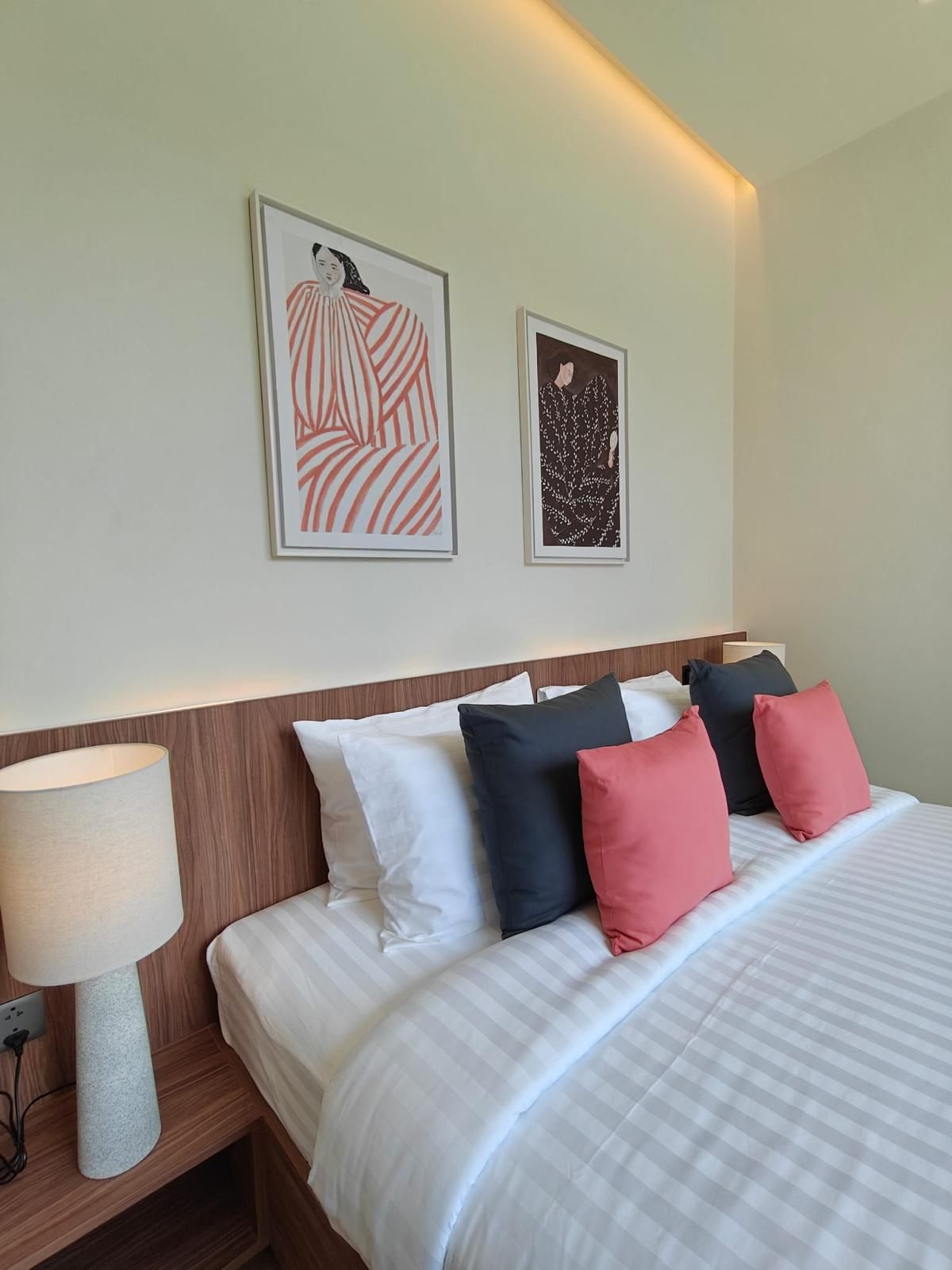Ein ordentlich bezogenes Bett mit weißer und gestreifter Bettwäsche, ein Holz-Kopfteil und eine Nachttischlampe. Über dem Bett hängen zwei gerahmte Kunstwerke.