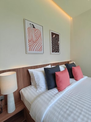 Ein ordentlich bezogenes Bett mit weißer und gestreifter Bettwäsche, ein Holz-Kopfteil und eine Nachttischlampe. Über dem Bett hängen zwei gerahmte Kunstwerke.