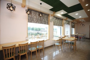 Un ristorante con tavoli e sedie in legno, ampie finestre con tende a motivi e lampade geometriche a sospensione.