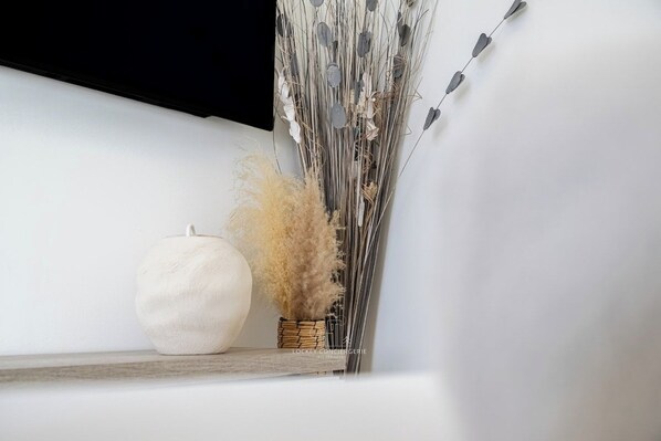 Un vase blanc contenant des plumes de jarre séchées, posé sur une étagère en bois devant un mur blanc.