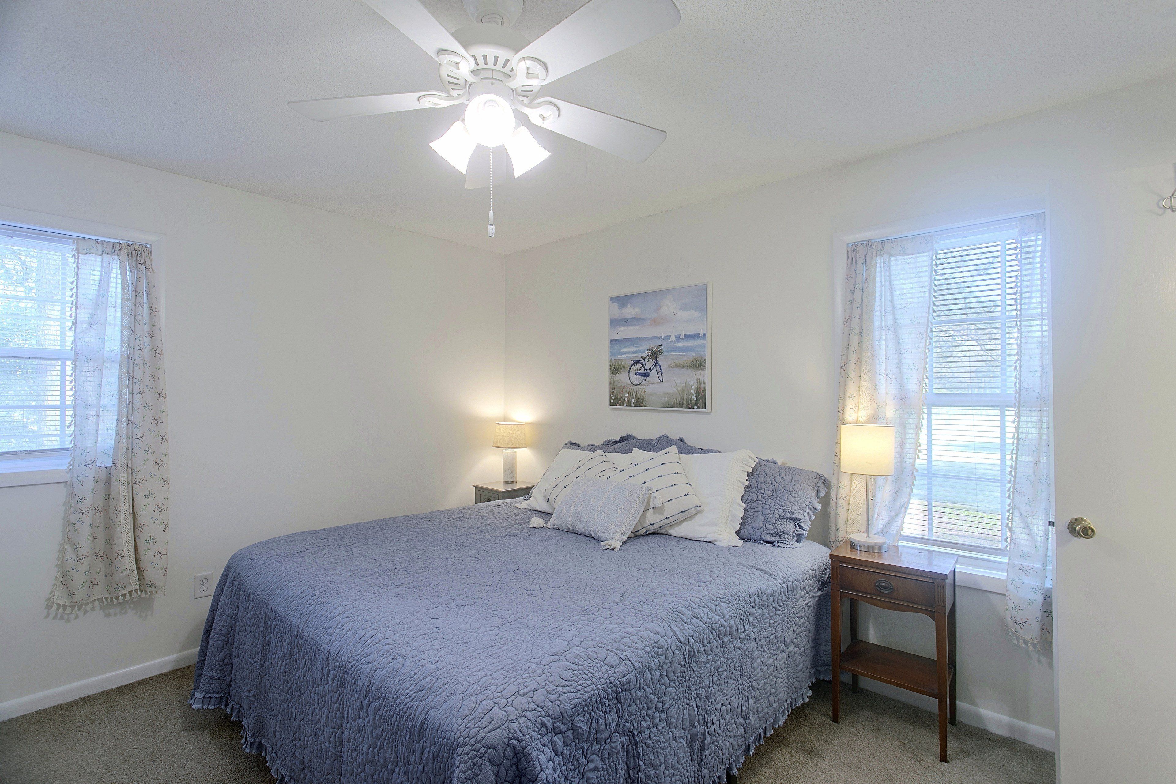 A bedroom with a bed, two bedside tables with lamps, a ceiling fan, and a window with curtains.