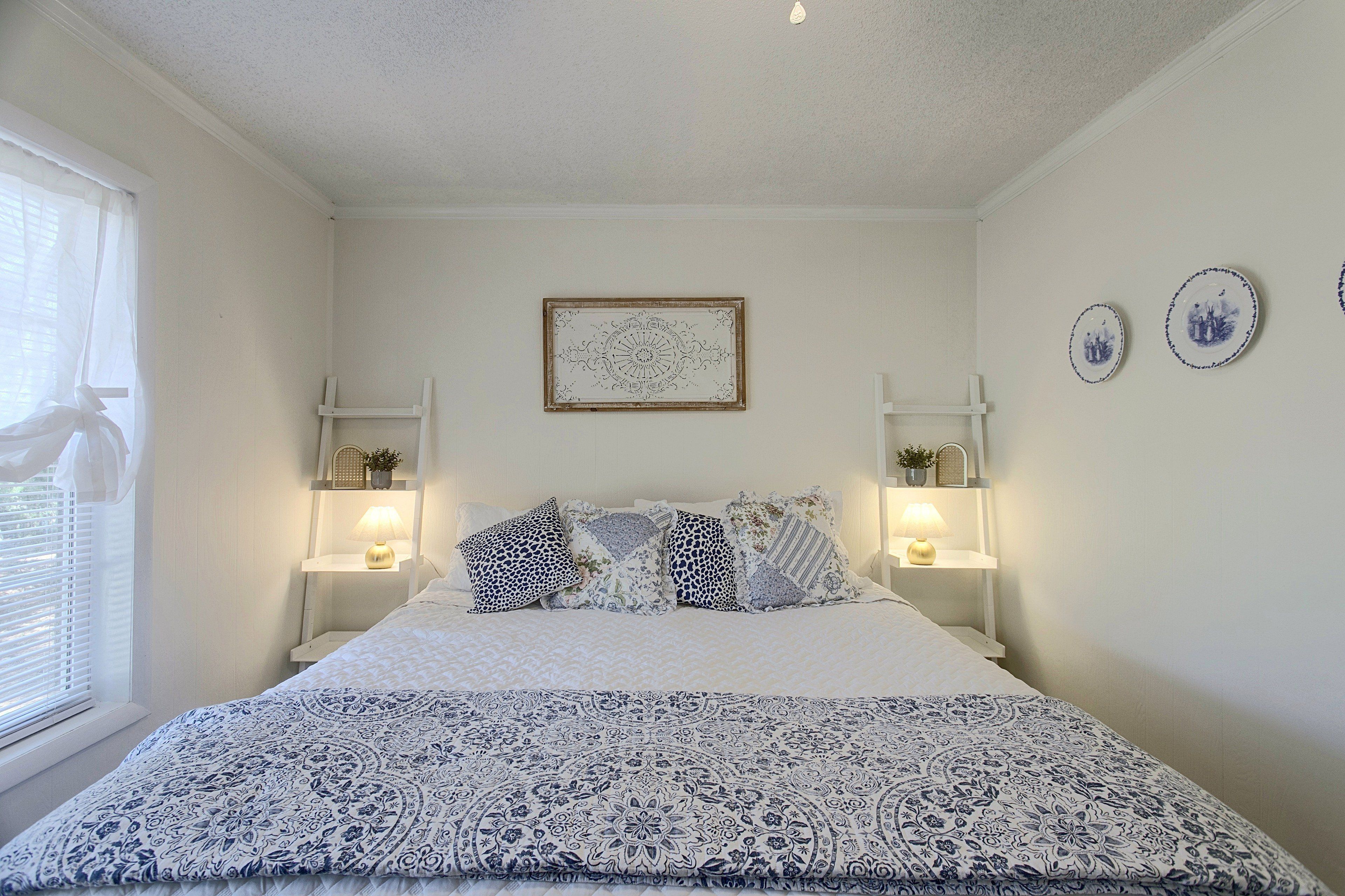 Quarto com cama, duas luminárias de cabeceira, janela com persianas e um quadro na parede.