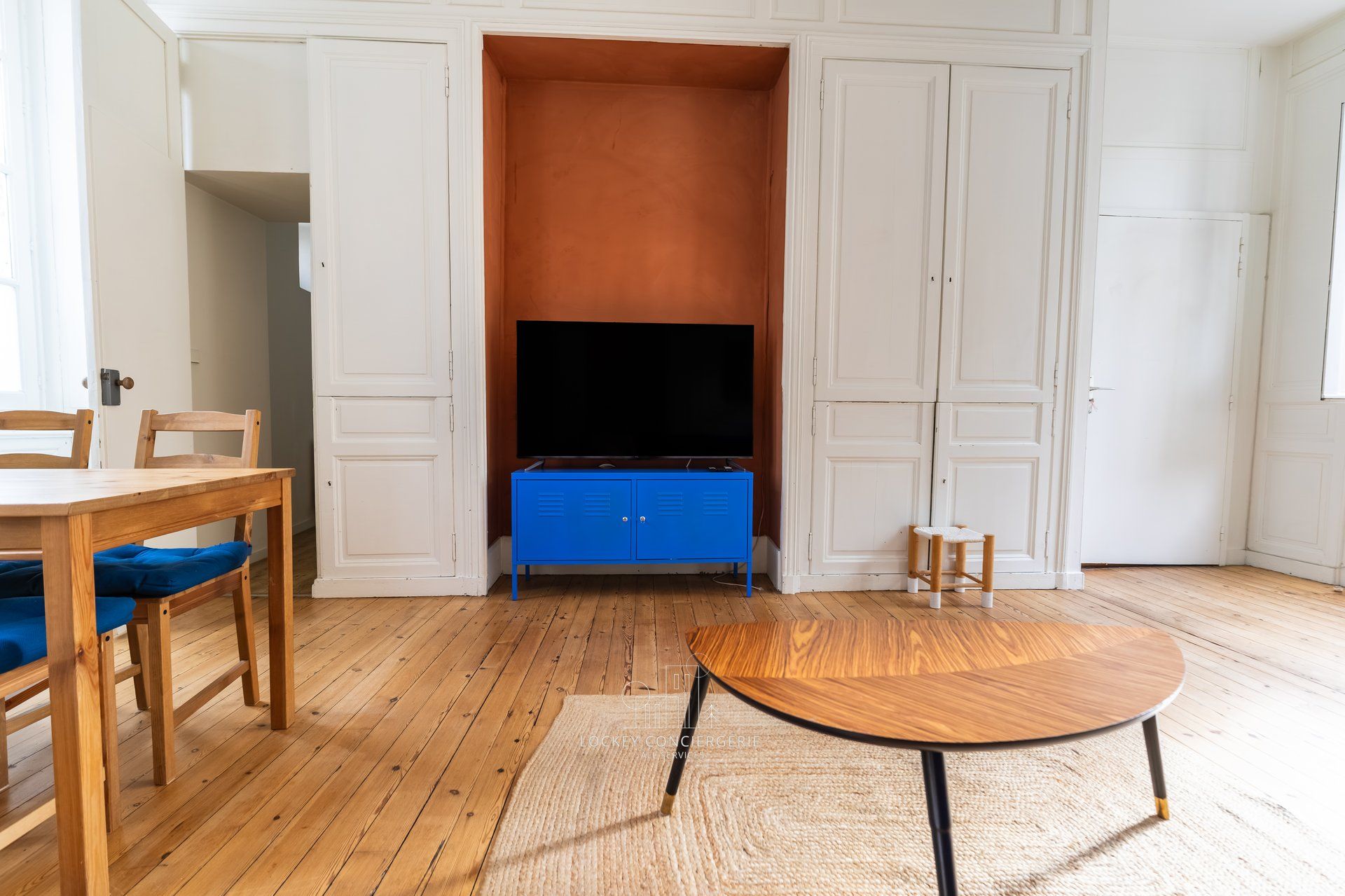 Una stanza con pavimento in legno, una TV su un mobiletto blu, un tavolo in legno e una sedia con cuscini blu.