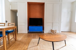 Una stanza con pavimento in legno, una TV su un mobiletto blu, un tavolo in legno e una sedia con cuscini blu.