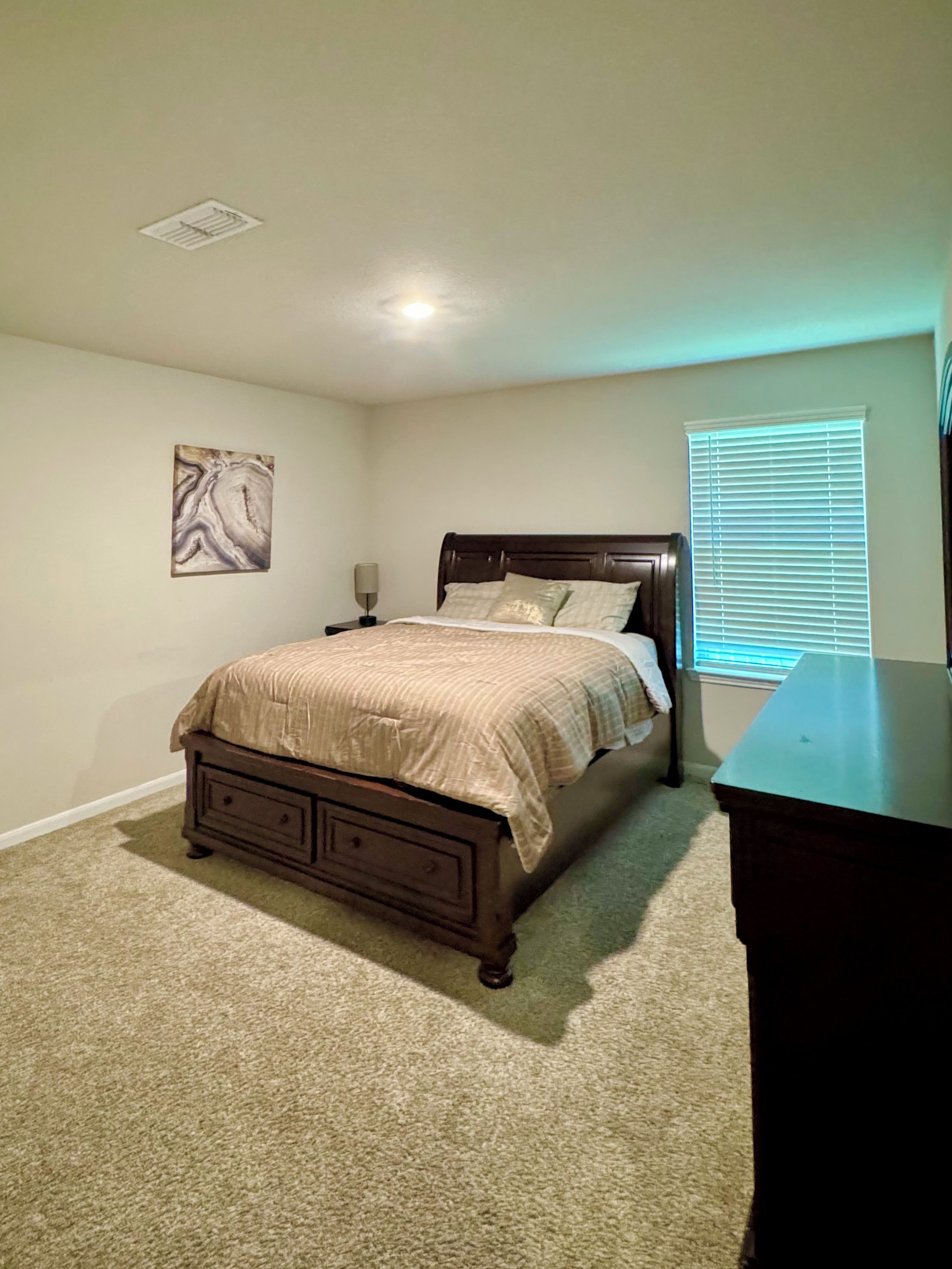 Une chambre à coucher comprenant un lit, une table de chevet, une lampe, une commode, une fenêtre avec des stores et une bouche d’aération au plafond.